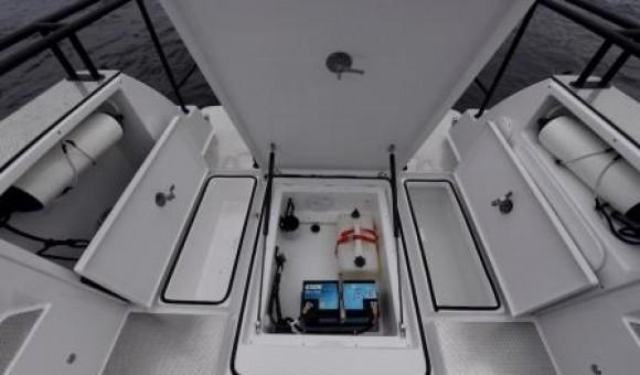 Open boat storage compartment featuring batteries, a fuel tank, and two side cabinets on a deck above water.